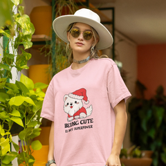 Person wearing a pink t-shirt with a cartoon dog in a Santa hat and text, standing in an outdoor setting with plants.