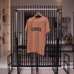 Brown t-shirt with 'COFFEE' text on a hanger against a black metal rack.