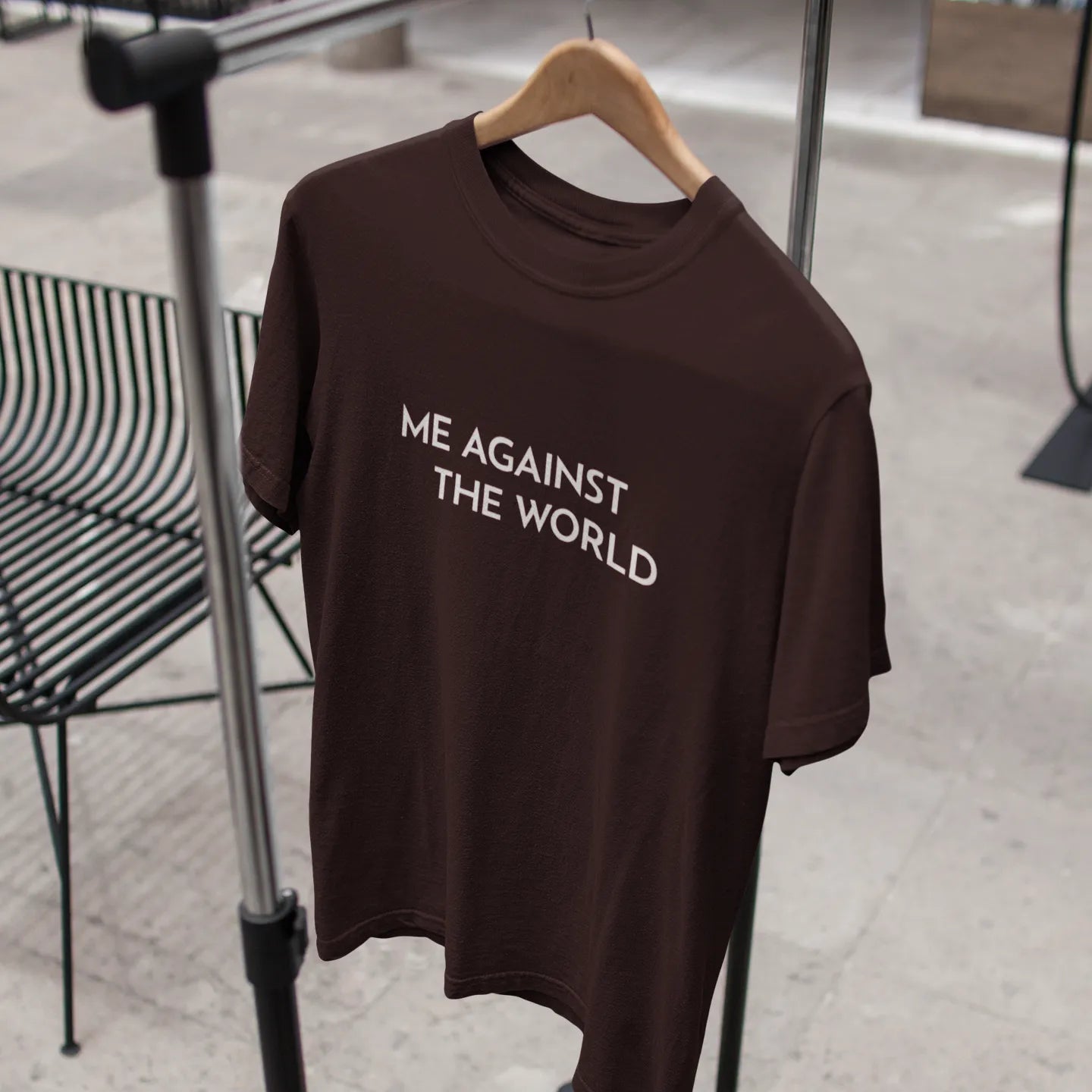 Brown t-shirt with 'ME AGAINST THE WORLD' text on a hanger against a blurred indoor background