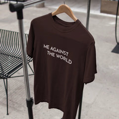 Brown t-shirt with 'ME AGAINST THE WORLD' text on a hanger against a blurred indoor background