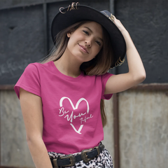 Young girl wearing a pink t-shirt with a heart design and text, standing against a textured wall.