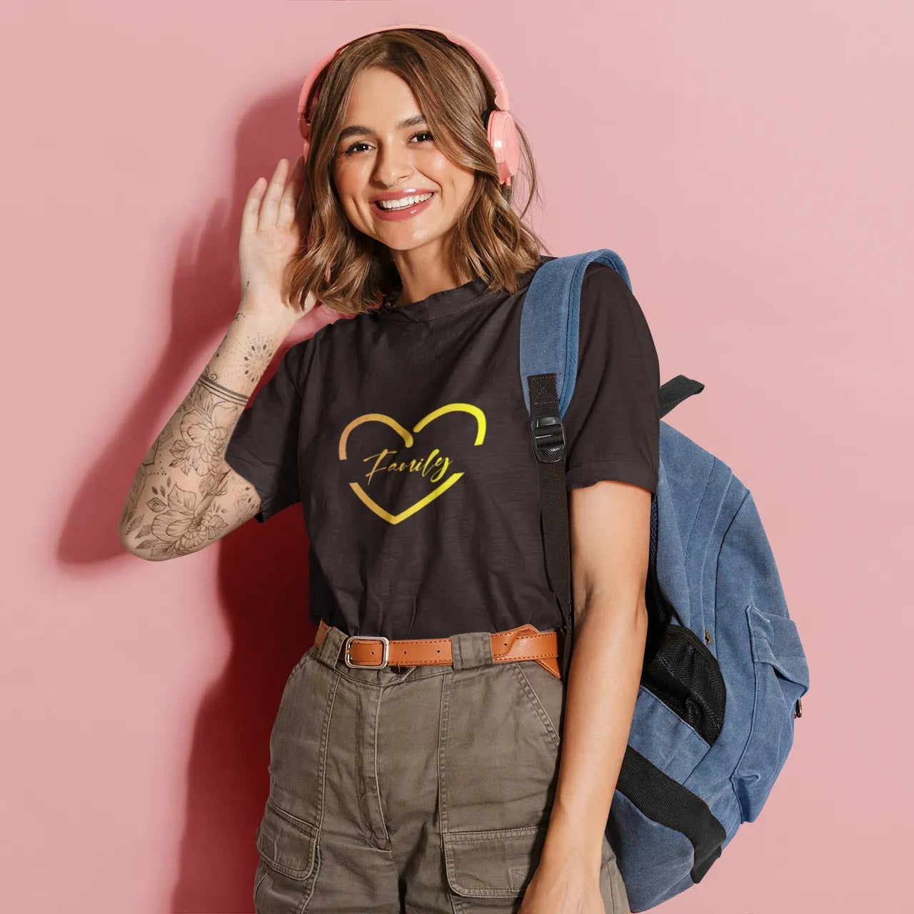 Woman wearing a black t-shirt with a yellow heart design, holding a blue backpack against a pink background
