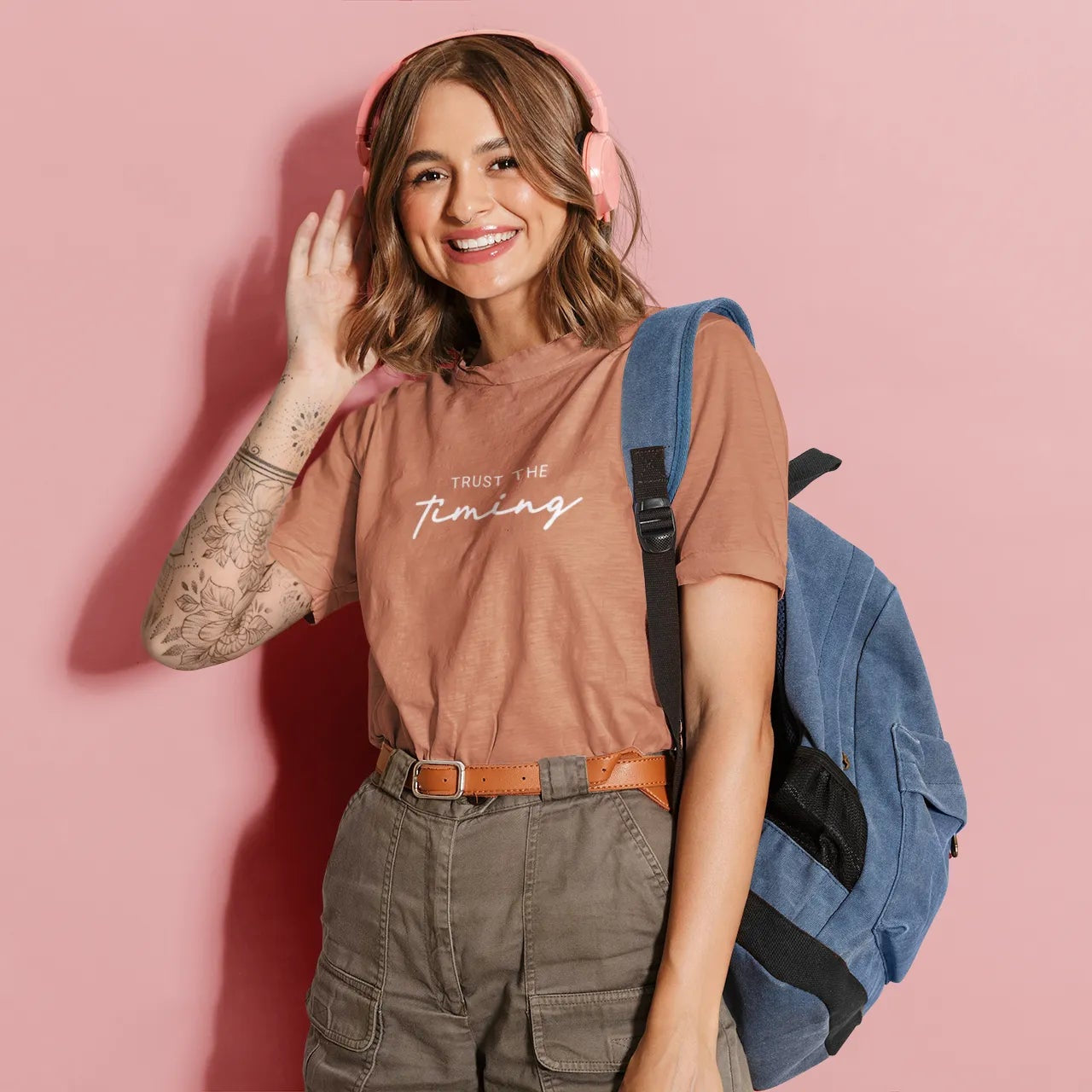 Woman wearing a brown t-shirt with text, green pants, and a blue backpack on a pink background