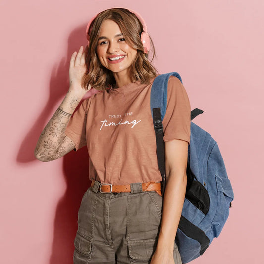 Woman wearing a brown t-shirt with text, green pants, and a blue backpack on a pink background
