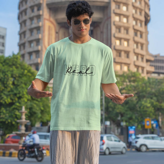 Person wearing a light green t-shirt with text, standing in an urban setting with buildings and trees in the background.