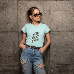 Woman wearing a light blue t-shirt with text, standing against a textured wall.