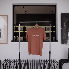 Brown t-shirt with 'GAME OVER' text hanging on a rack in a modern interior setting.