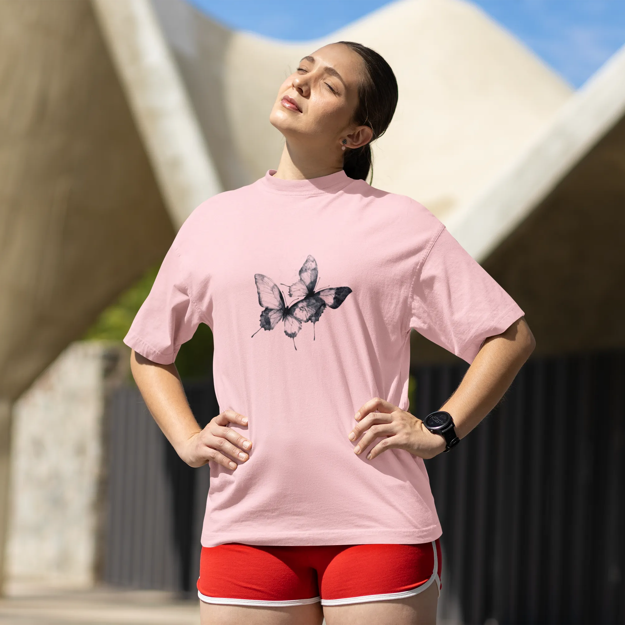 Woman wearing a pink t-shirt with a butterfly design and red shorts outdoors.
