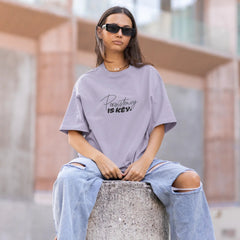 Person wearing a gray oversized t-shirt with text and ripped jeans sitting on a concrete block.