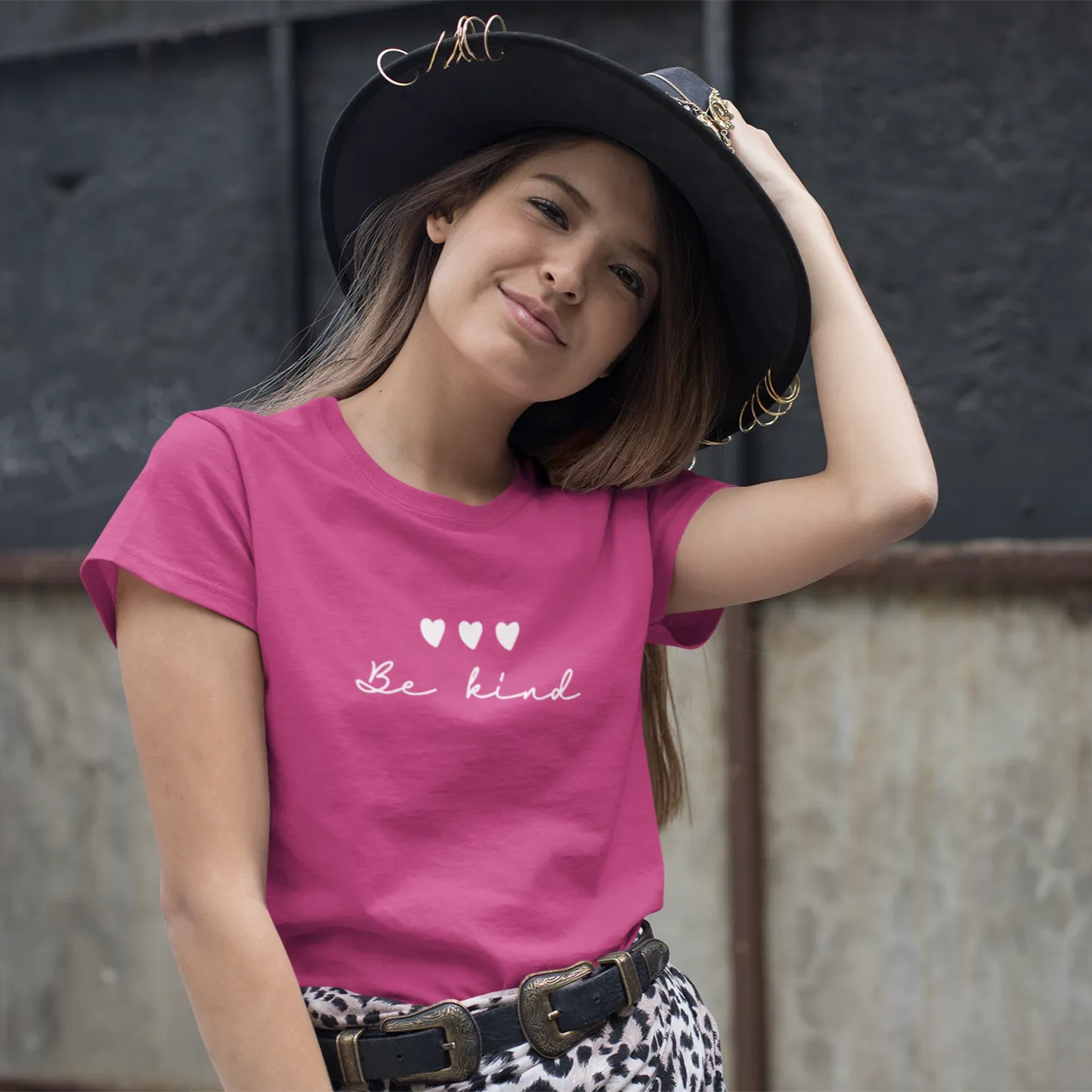 Woman wearing a pink t-shirt with 'Be kind' text and hearts, standing against a textured wall.