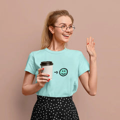 Woman wearing a light blue t-shirt with a smiley face design, holding a coffee cup, and waving on a beige background.