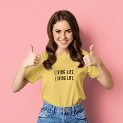 Woman wearing a yellow t-shirt with 'LIVING LIFE LOVING LIFE' text, giving thumbs up on a pink background