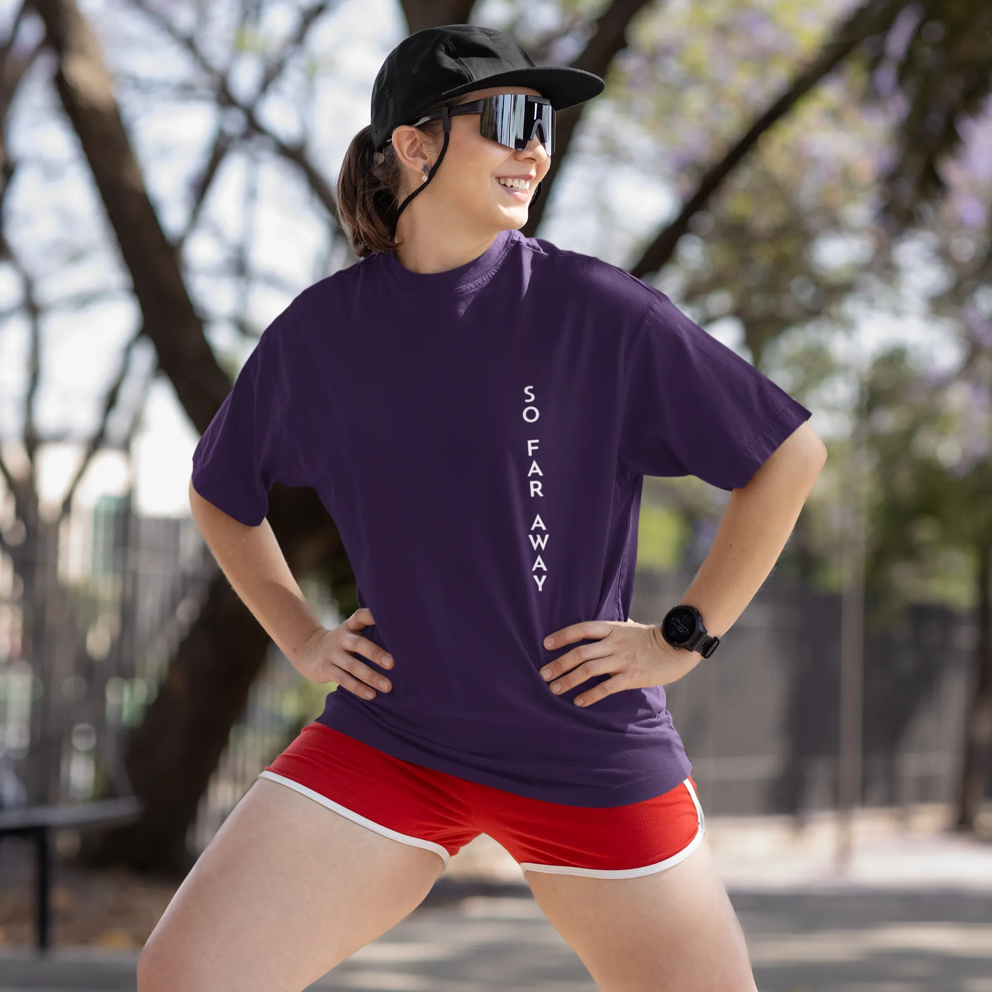 Woman wearing a purple t-shirt and red shorts standing outdoors with trees in the background