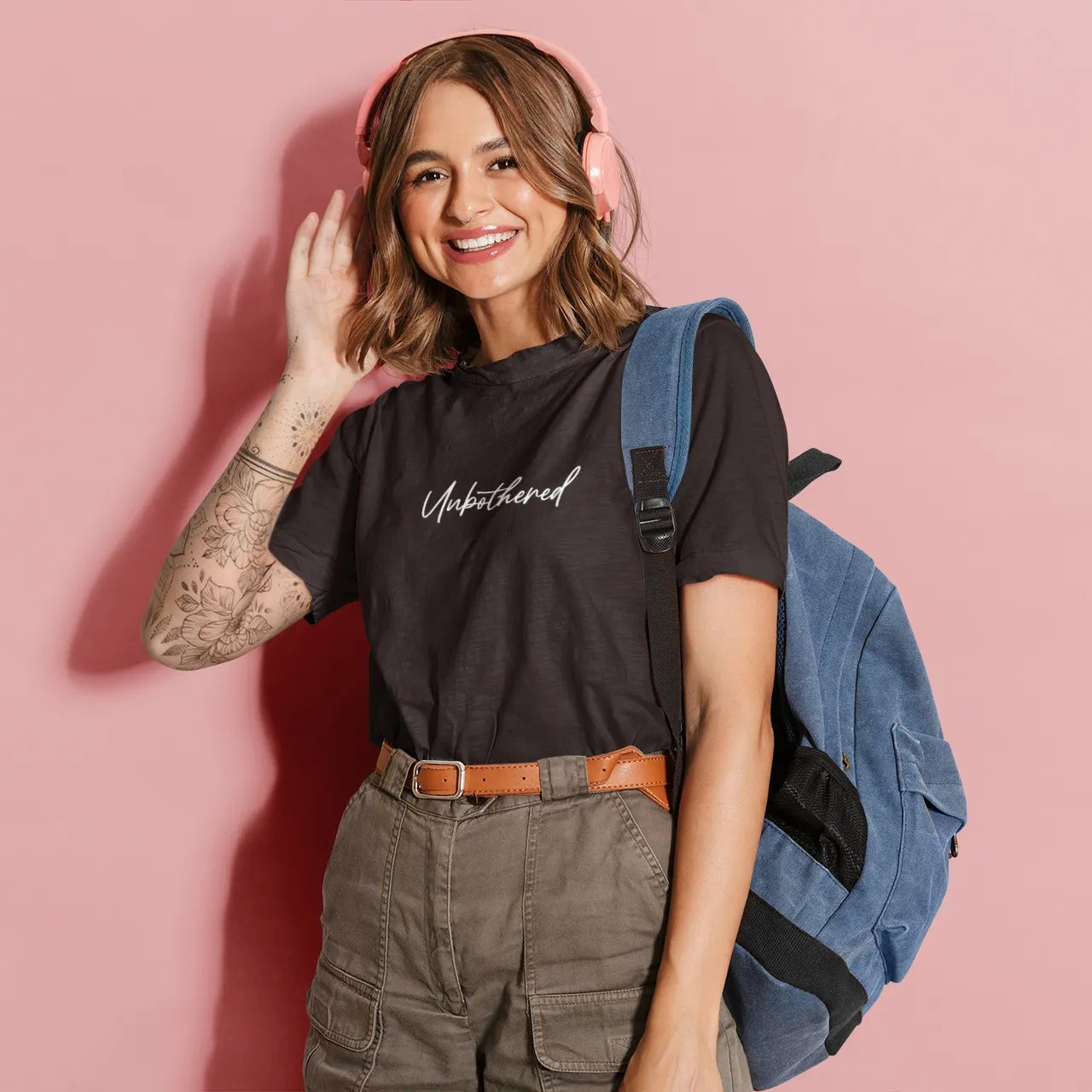 Woman wearing a black t-shirt with text, green cargo shorts, and a blue backpack on a pink background