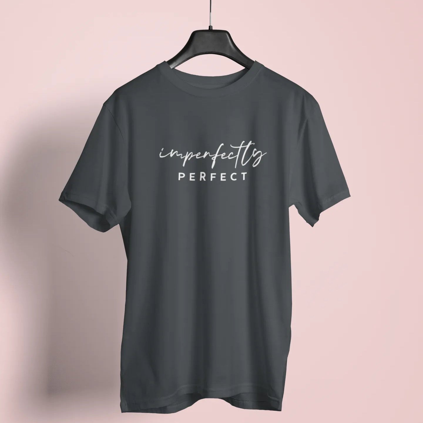 Dark gray t-shirt with 'imperfectly perfect' text on a hanger against a pink background