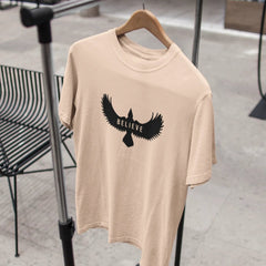 Beige t-shirt with an eagle graphic and 'BELIEVE' text on a hanger.