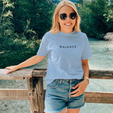 Woman wearing a light blue t-shirt with 'BALANCE' text, denim shorts, and sunglasses, standing by a wooden railing with a scenic background.