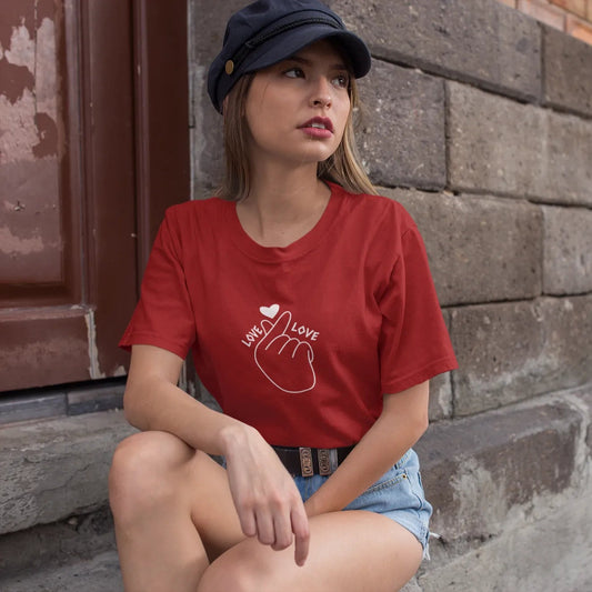 Woman wearing a red t-shirt with a heart design sitting against a brick wall.