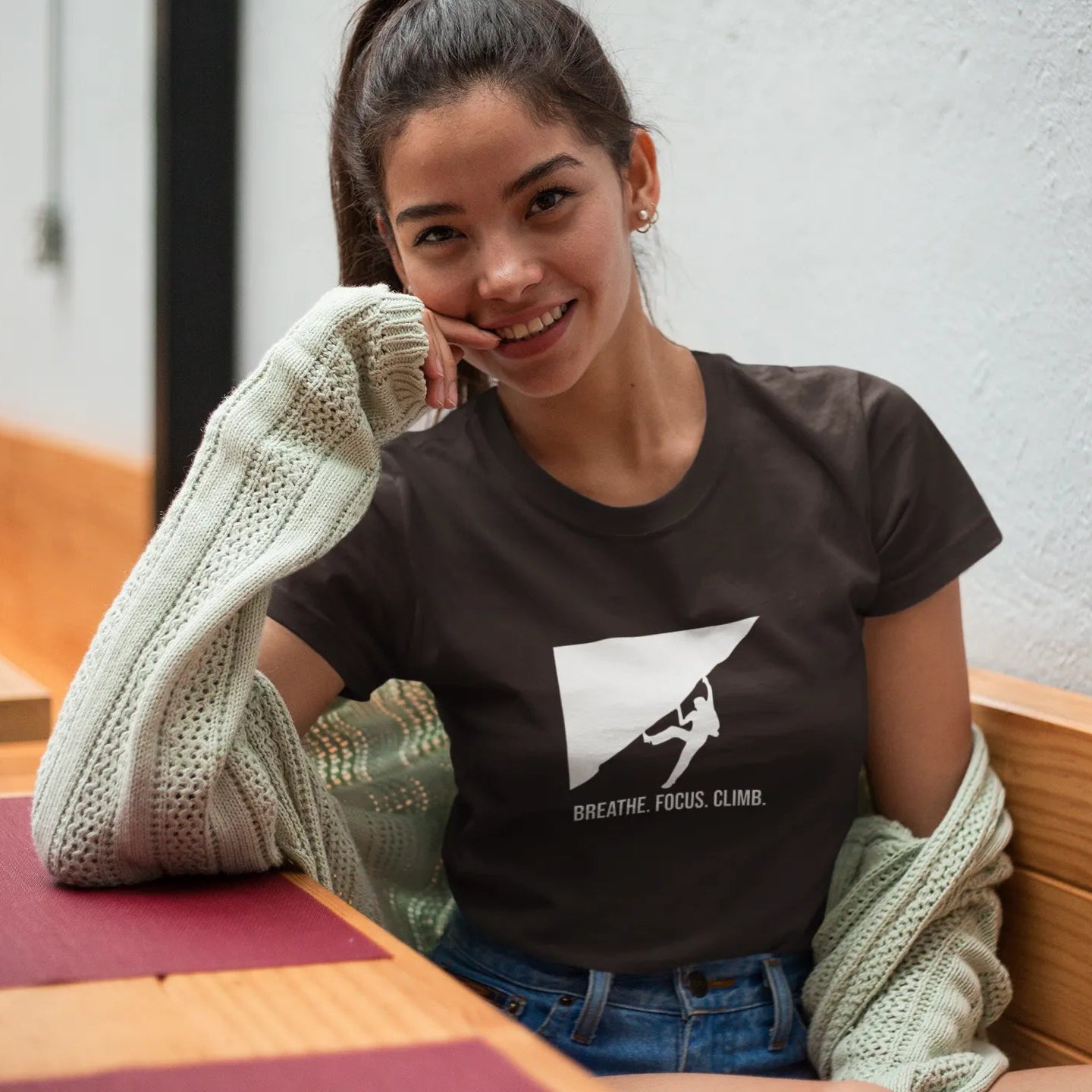 Woman wearing a t-shirt with a climbing graphic and text, sitting indoors.
