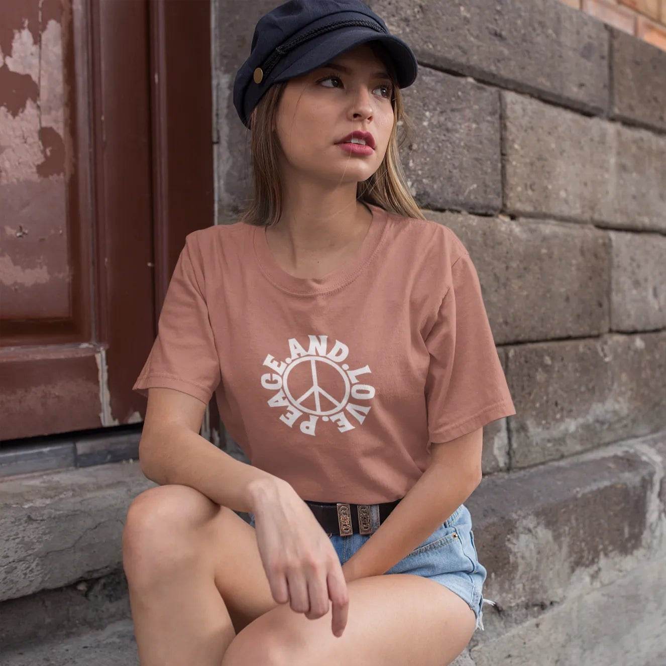 Woman wearing a pink t-shirt with text and a peace symbol, sitting against a brick wall.