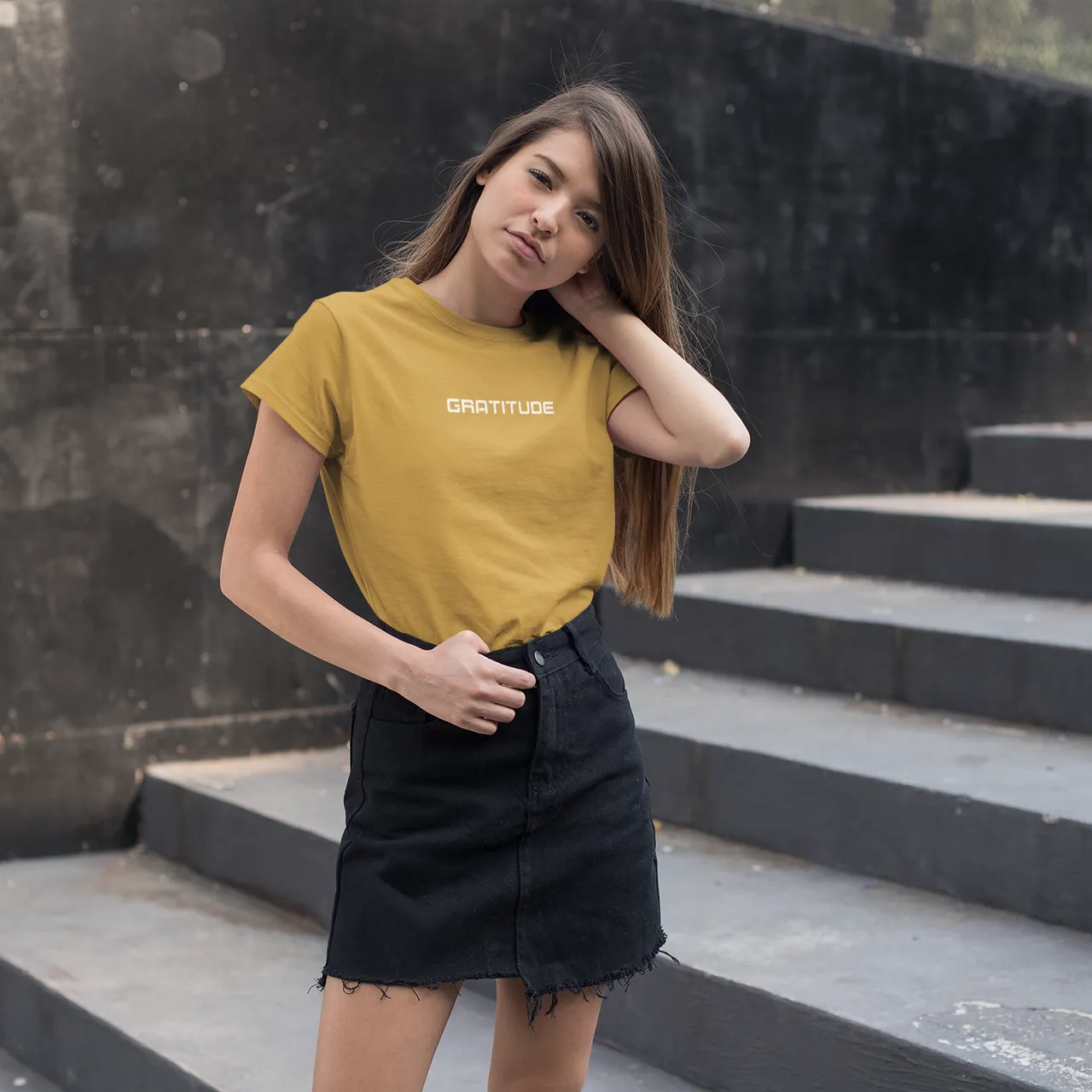 Woman wearing a mustard yellow t-shirt with text and black skirt standing on steps.