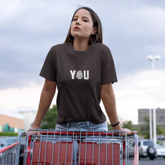 Woman wearing a brown t-shirt with 'YOU' printed on it, standing next to a shopping cart.