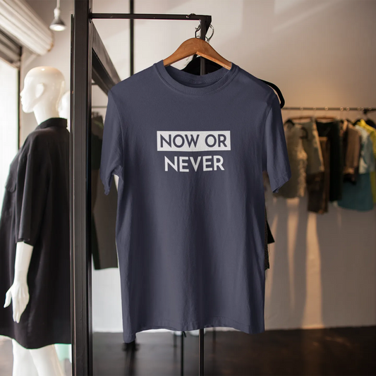 Navy blue t-shirt with 'NOW OR NEVER' text on a hanger in a clothing store.