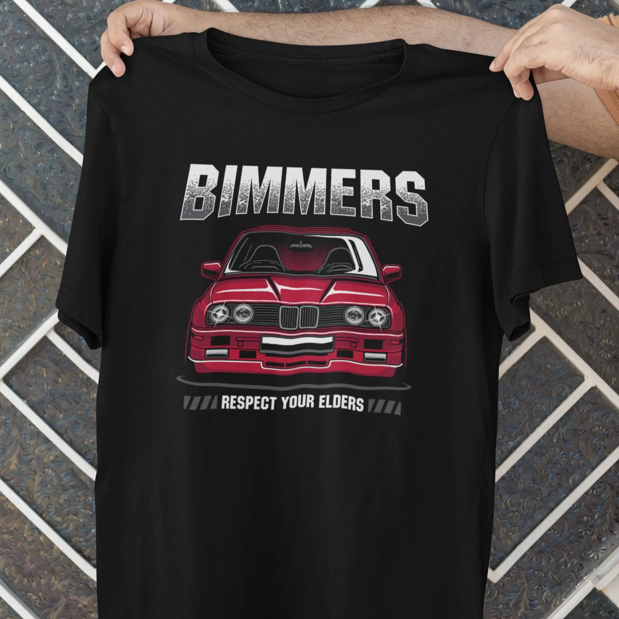 Black t-shirt with a red car graphic and 'Bimmers' text held by a person against a tiled wall.