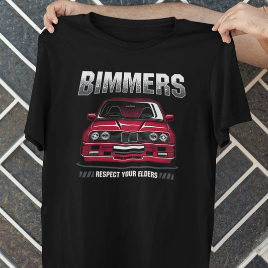 Black t-shirt with a red car graphic and 'Bimmers' text held by a person against a tiled wall.