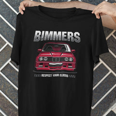 Black t-shirt with a red car graphic and 'Bimmers' text held by a person against a tiled wall.