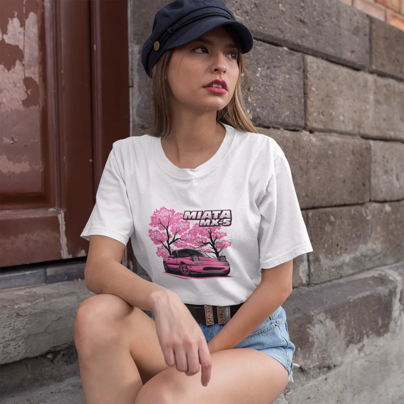 Woman wearing a white t-shirt with a car graphic sitting against a brick wall.