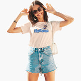 Woman wearing a light pink t-shirt with a graphic design and denim shorts, posing against a white background.