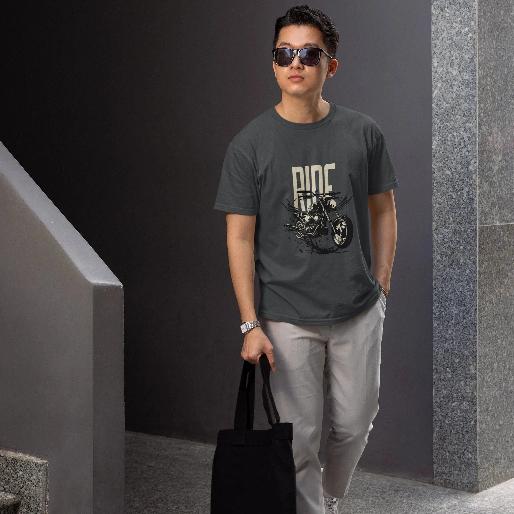 Man wearing sunglasses and a dark t-shirt with a graphic design, holding a black tote bag, standing against a gray wall.