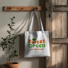 White tote bag with 'Sweet Green' text hanging on a wooden hook next to a plant.