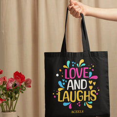 Black tote bag with colorful text 'LOVE AND LAUGHS' held by a hand on a beige curtain background.