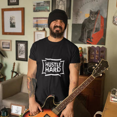 Man holding a guitar wearing a 'Hustle Hard' t-shirt in a room with various decorations.