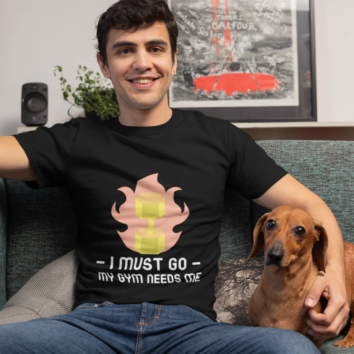 Man sitting on a couch with a dog, wearing a black t-shirt with a graphic design and text.