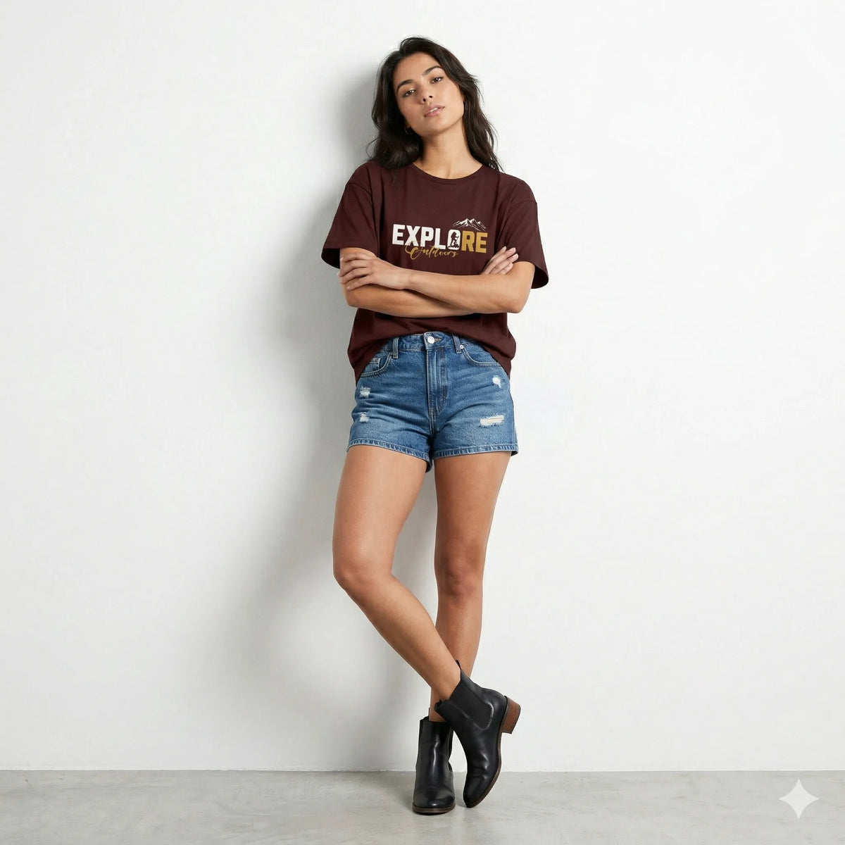 Woman wearing a brown 'EXPLORE' t-shirt, blue denim shorts, and black ankle boots against a white background.