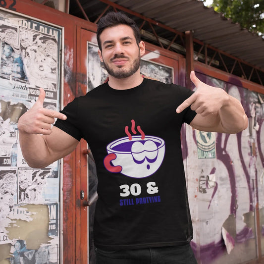Man wearing a black t-shirt with a graphic design, pointing at it in front of a wall with posters.