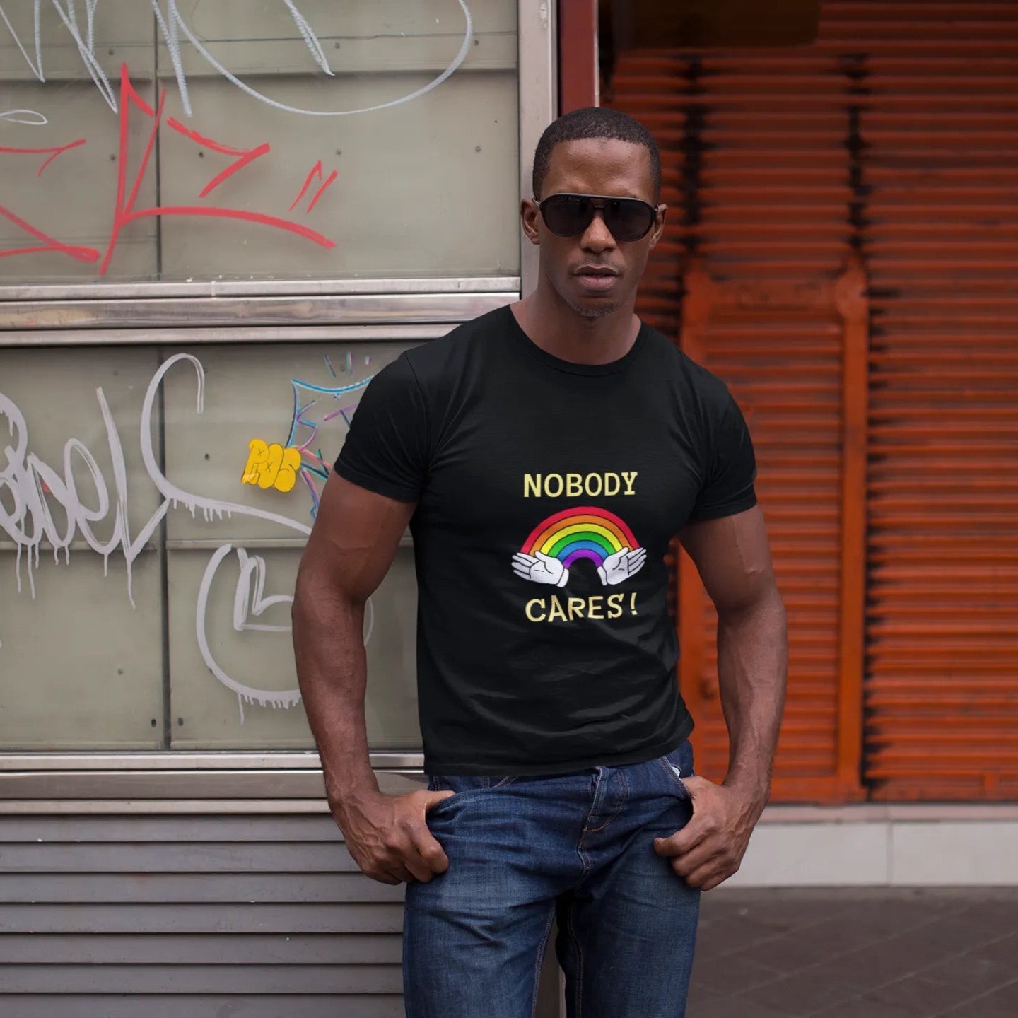 Man wearing a black t-shirt with 'NOBODY CARES!' text and rainbow design, standing in front of a graffiti-covered wall.