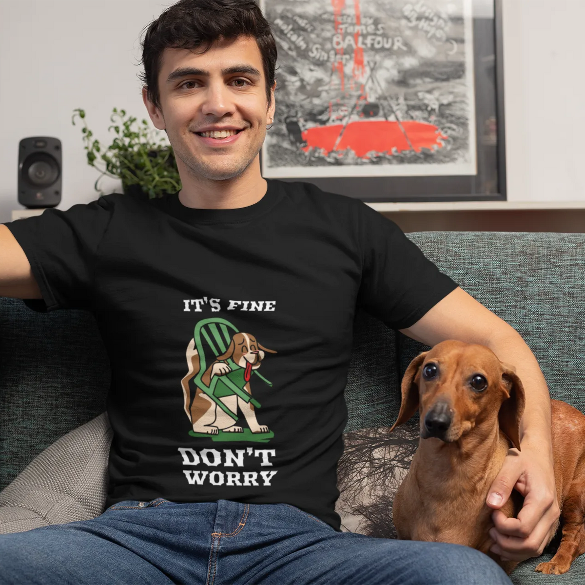 Man sitting on a couch with a dog, wearing a black t-shirt with a graphic and text.