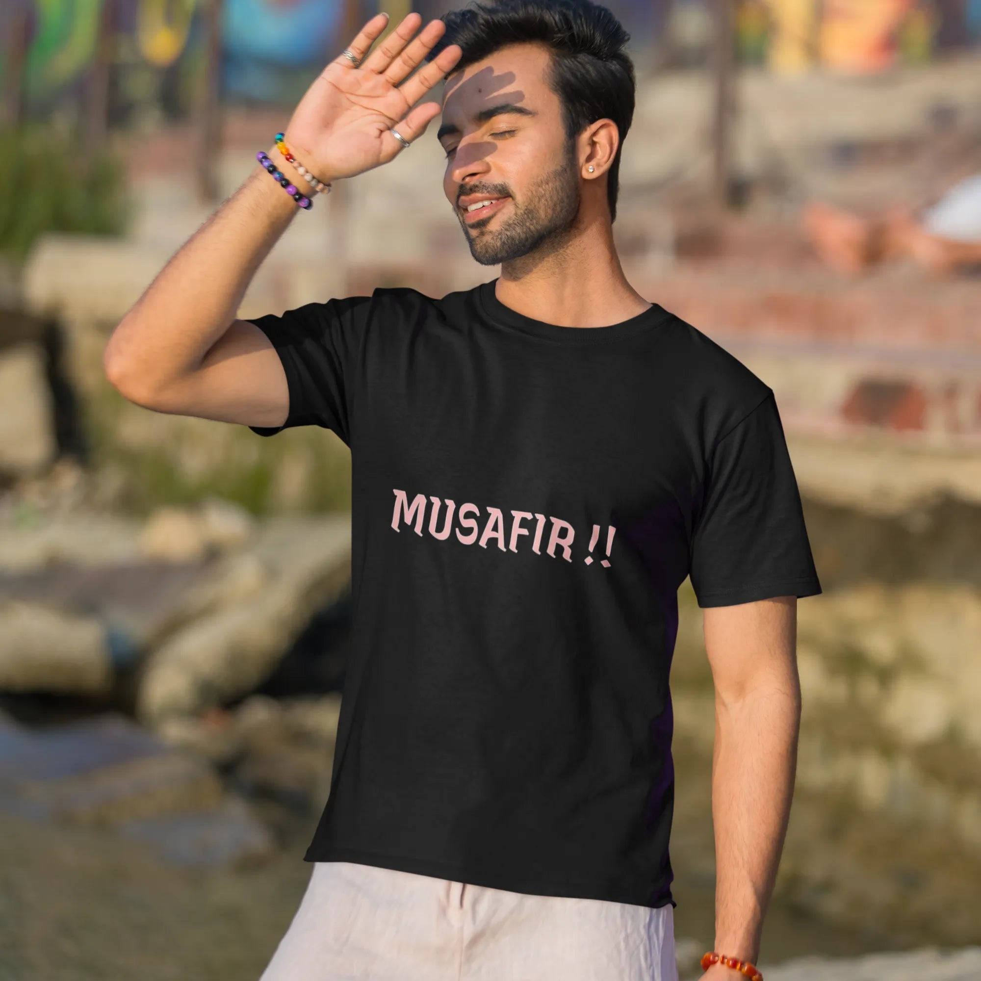 Man wearing a black t-shirt with 'MUSAFIR!!' text, standing outdoors with a colorful mural in the background.