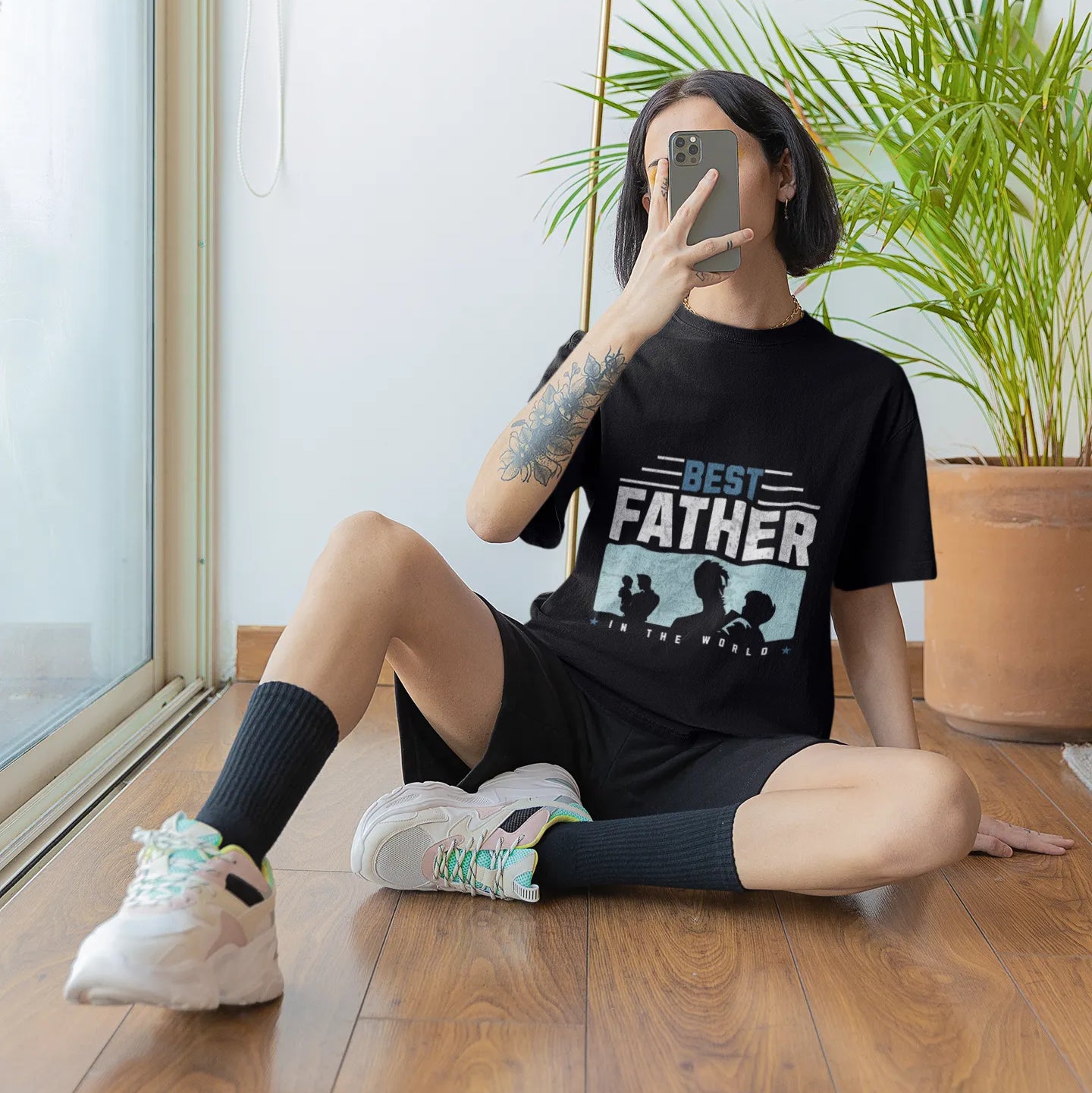 Person sitting on the floor wearing a black t-shirt with 'Best Father' graphic, taking a mirror selfie.