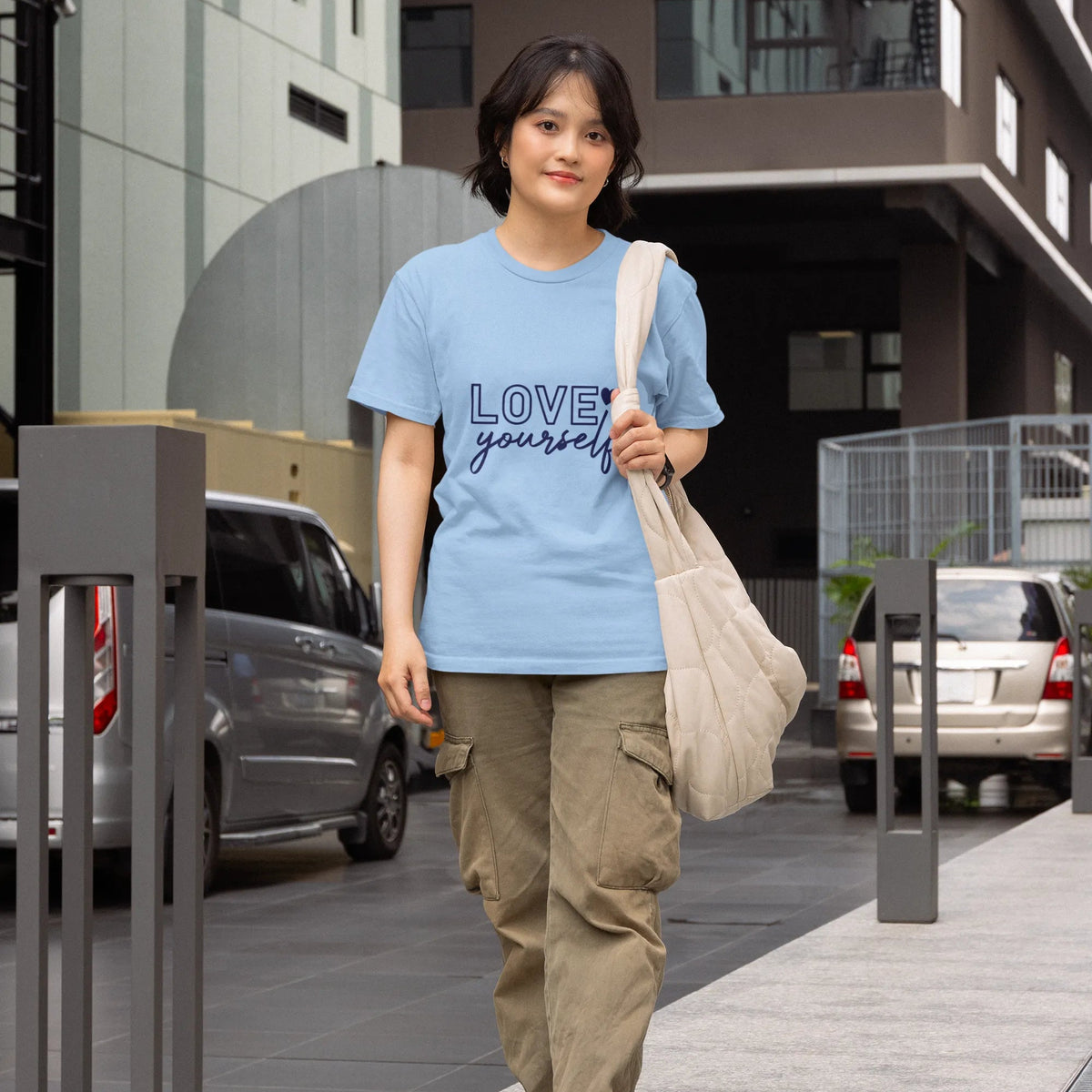 Person wearing a blue t-shirt with text, walking on a sidewalk with buildings and cars in the background.