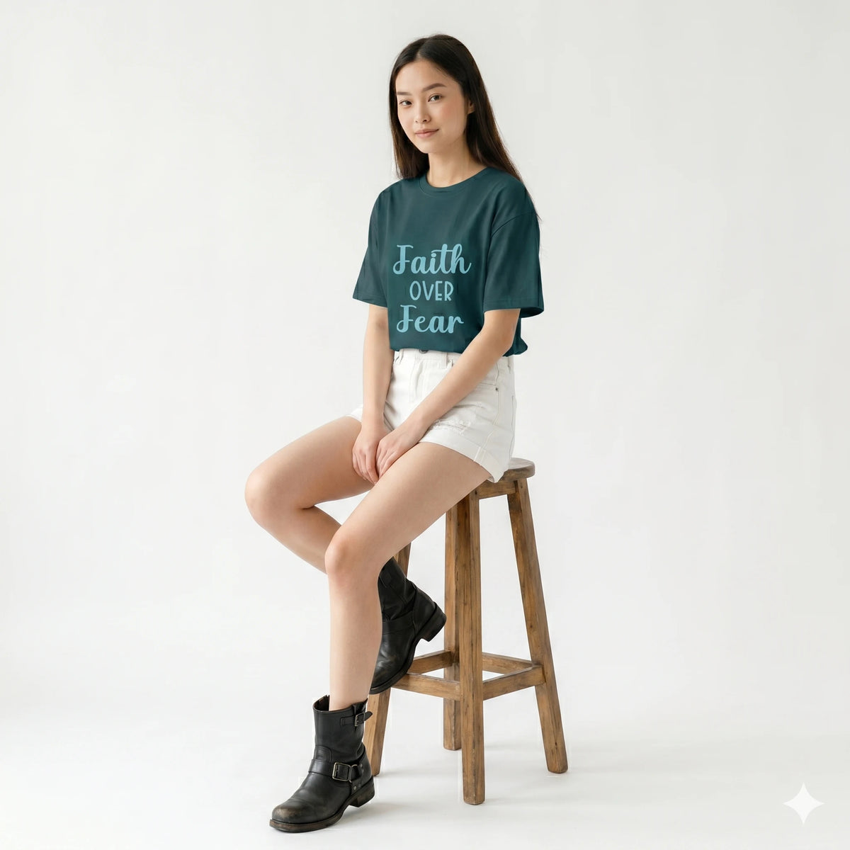 Person wearing a teal t-shirt with 'Faith Over Fear' text, sitting on a wooden stool against a white background.