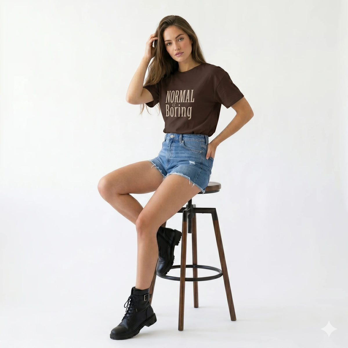 Woman sitting on a stool wearing a brown t-shirt with text and denim shorts on a white background