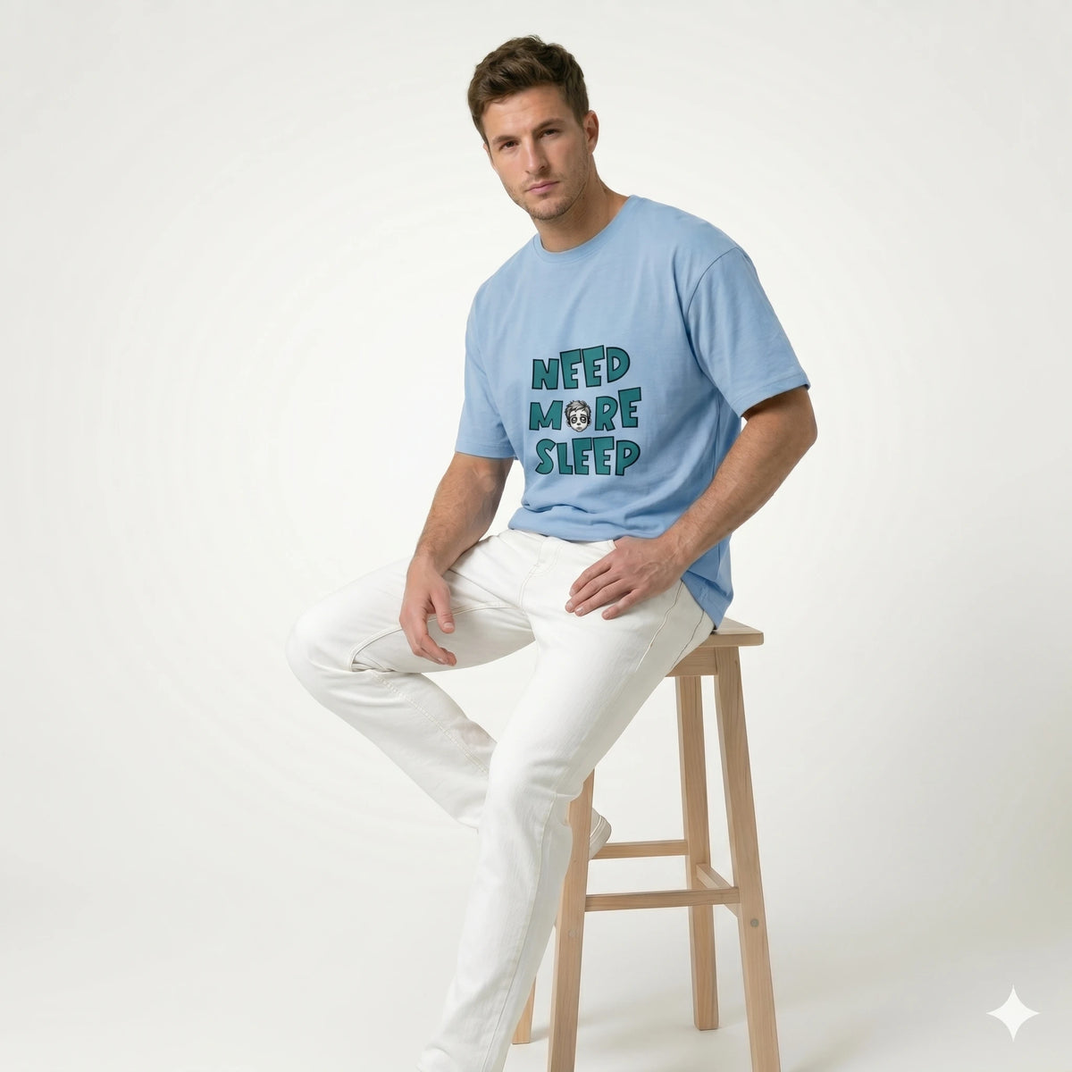 Man sitting on a stool wearing a light blue t-shirt with text and white pants on a plain background