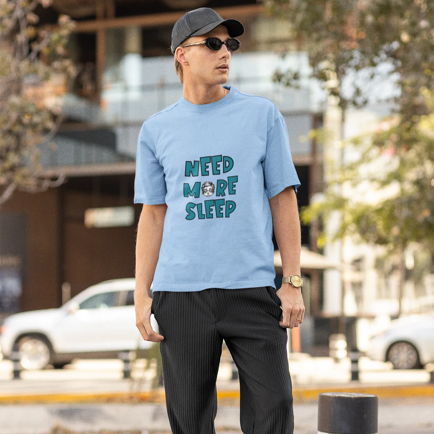 Man wearing a light blue t-shirt with text, dark pants, and cap on an urban street.