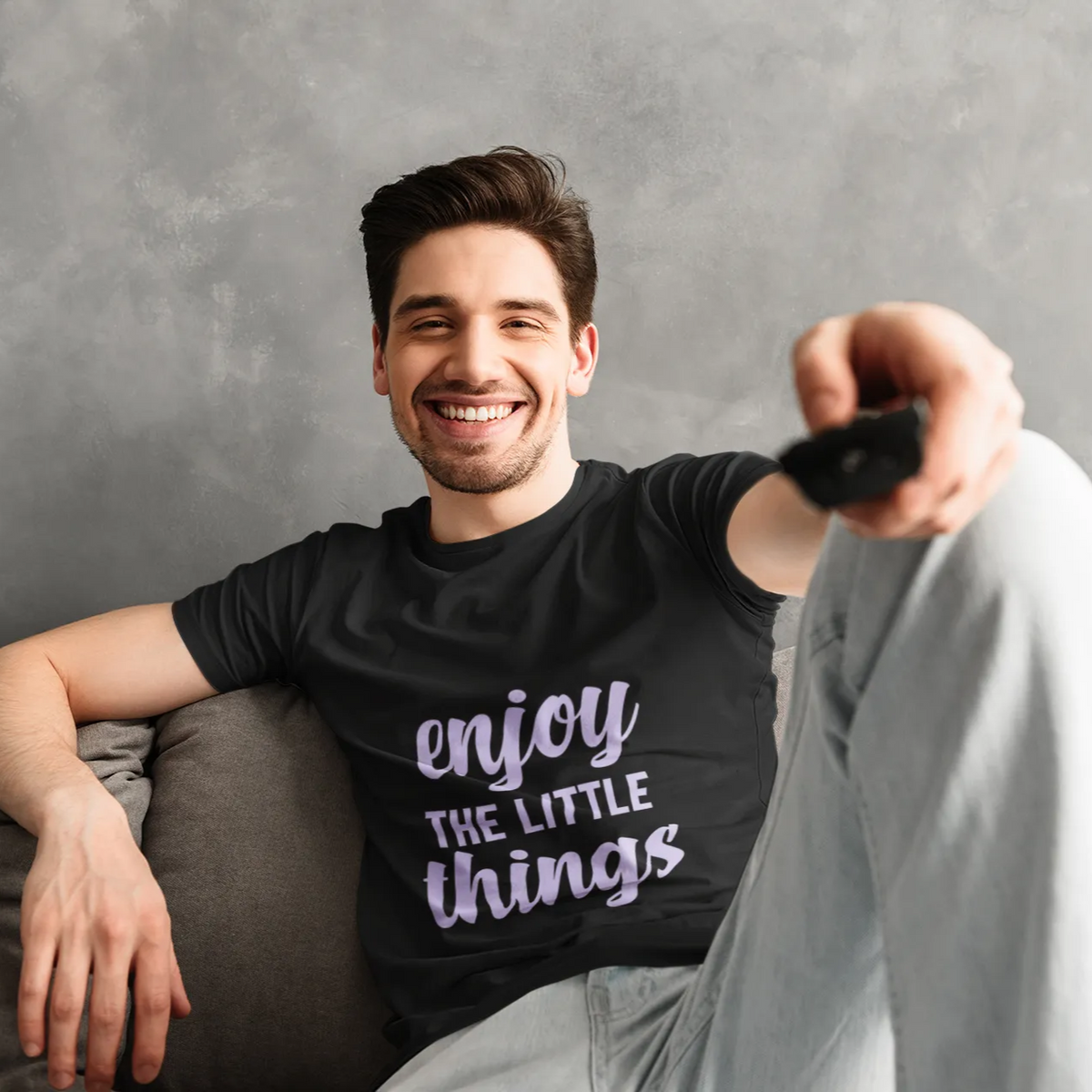 Man sitting on a couch wearing a black t-shirt with 'enjoy the little things' text, holding a remote control.
