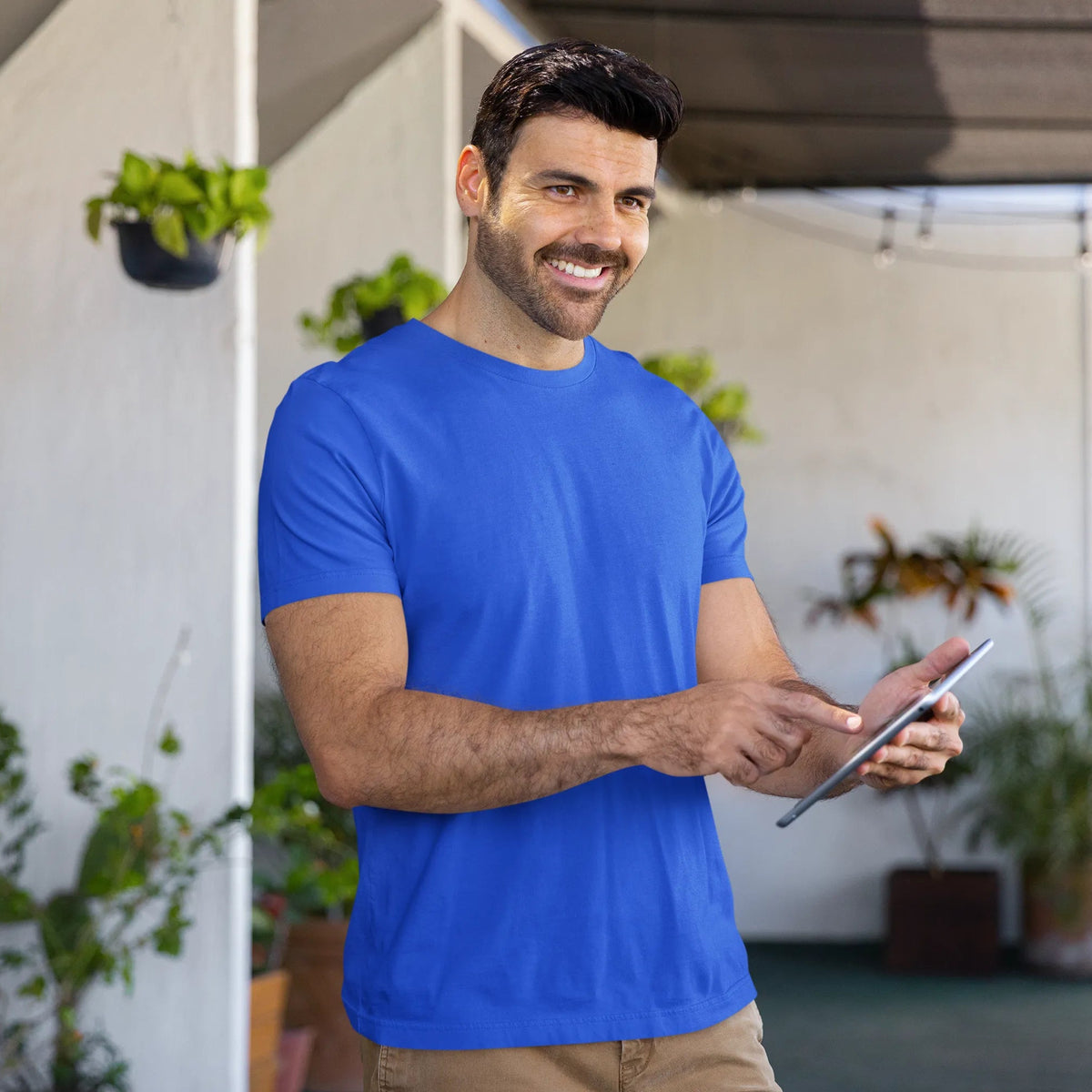 Man in a blue shirt holding a tablet outdoors with plants in the background